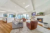A spacious, modern living room with a high, white, wood-beamed ceiling. It features a cozy seating area with a gray sofa, two brown leather chairs, a fireplace, and a flat-screen TV. Large windows provide natural light and views of Shelter Cove, while the open kitchen is visible in the background.