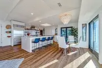 A modern, open-concept kitchen and dining area in Shelter Cove boasts large windows that bathe the space in natural light. The kitchen features a white island with bar stools, built-in appliances, and a dining table with white chairs. Wooden floors and contemporary decor complete the room's elegant design.