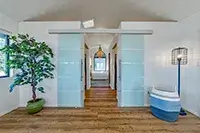 A modern hallway with light blue sliding doors on both sides evokes the serene ambiance of Shelter Cove. A large plant in a green pot is on the left, and a white chair with blue cushions is on the right. Wooden flooring is underfoot, and a pendant light hangs from the ceiling, leading to a room with a large window.