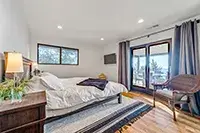 A bright bedroom in Shelter Cove with a large bed covered in white bedding, flanked by a wooden nightstand with a lamp. The room features wooden floors, a window with a view, and a glass door leading to an outdoor area. There's a cushioned chair by the door and framed artwork on the wall.