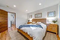 A cozy bedroom in Shelter Cove features a wooden bed frame, white bedding, and a blue throw blanket. Two wooden nightstands with lamps and decorative items are on either side of the bed. The room has hardwood floors, rugs, and framed pictures on the wall, with an open door on the left.