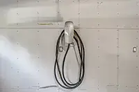 A silver electric vehicle charging unit is mounted on a white wall, blending seamlessly with the clean, industrial look of the garage. The black charging cable is neatly coiled and hanging from the unit, while visible screws and slight shadows add to the aesthetic.