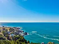 Coastal landscape featuring a clear, blue sky and a vast, calm ocean. The shoreline of Shelter Cove is rocky with scattered greenery, and waves gently crash against the rocks. The horizon is visible, creating a tranquil and picturesque scene.