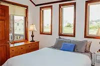 A cozy bedroom in Shelter Cove features a large bed with white and gray bedding and a blue accent pillow. A wooden dresser with a lamp is placed next to the bed. Three windows with views of houses and greenery allow natural light to illuminate the room.