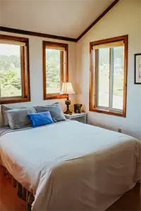 A cozy bedroom in Shelter Cove features a neatly made bed with white linens and a blue accent pillow. Three large windows with wooden frames allow natural light to fill the space. A bedside table adorned with a lamp and decorative items is situated next to the bed, completing this inviting retreat.