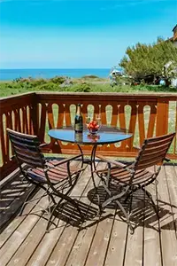 A wooden deck with a round glass table and two wooden chairs overlooks a scenic view of the ocean at Shelter Cove. On the table are a bottle of wine, two wine glasses, and a bowl of strawberries. Trees and greenery are visible in the background under a clear blue sky.