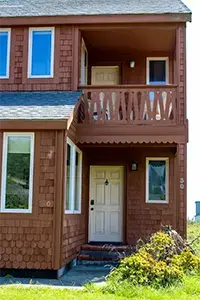 A charming two-story house in Shelter Cove with brown shingles and white trim. The front door is on the lower level with wooden steps leading to it. The upper level has a small balcony with a decorative railing. Several windows reflect the blue sky and greenery surrounding the home.