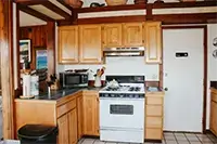 A cozy kitchen in Shelter Cove features wooden cabinets and countertops. A white stove and oven is centered beneath a stainless steel range hood. To the left, there's a microwave, kitchen utensils, and a trash bin. Baskets are displayed on top of the cabinets.