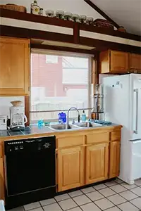 A kitchen in Shelter Cove with wooden cabinets, a double sink under a large window, a black dishwasher, and a white refrigerator. The countertop holds several appliances, including a coffee maker and blender. Shelves above the window display glass containers.