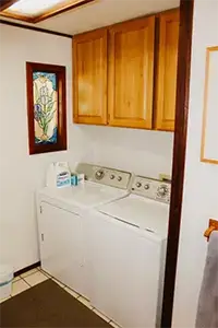 A small laundry room with a white washing machine and dryer side by side beneath wooden cabinets exudes the charm of Shelter Cove. On the left wall, a decorative stained glass window adds a touch of elegance. A container of detergent sits on top of the washing machine, while the tiled floor completes the cozy setup.