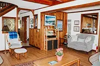 Cozy living room in Shelter Cove with wooden paneling and a mix of modern and rustic decor. It features a cushioned armchair with a blue throw blanket, a potted plant on a wooden coffee table, wall art, and a beige loveseat. The room has wooden beams and ample natural light.