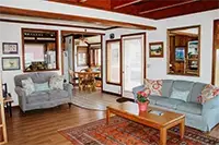 A cozy living room in Shelter Cove features two cushioned sofas with decorative pillows, a wooden coffee table with a potted plant, and a patterned rug on wooden flooring. The room has wooden ceiling beams and large windows offering natural light, leading to a dining area in the background.