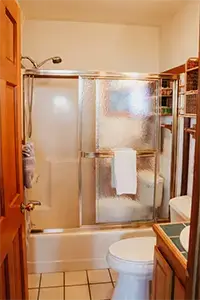 A compact bathroom in Shelter Cove features a shower with frosted sliding glass doors. A white towel hangs on the shower door. The room includes a white toilet and a sink with a mirror above. Wooden shelves on the right hold various toiletries.