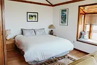 A cozy Shelter Cove bedroom with a large bed covered in white bedding and flanked by two wooden nightstands with lamps. The room features wooden accents, a framed picture on the wall, and a sunlit window seat with cushions. The floor is finished with a woven rug.