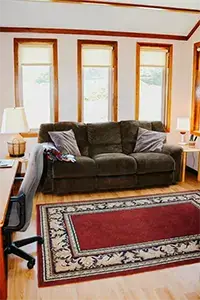 A cozy living room at Shelter Cove features a dark brown couch with gray pillows, situated against a wall with four windows. The room is adorned with a red and beige decorative rug on a wooden floor. A desk with a chair and a lamp sits on the left side.