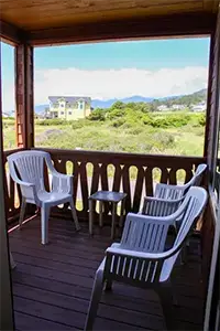 A view from a wooden deck with three white plastic chairs and a small table. The deck overlooks the grassy field of Shelter Cove, with a yellow house and mountains in the background under a partly cloudy sky.