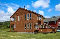A two-story wooden house with many windows stands on a grassy lawn at Shelter Cove, set against green hills and a bright blue sky with scattered clouds. The home features an outdoor deck on the ground level and a covered balcony on the upper level.
