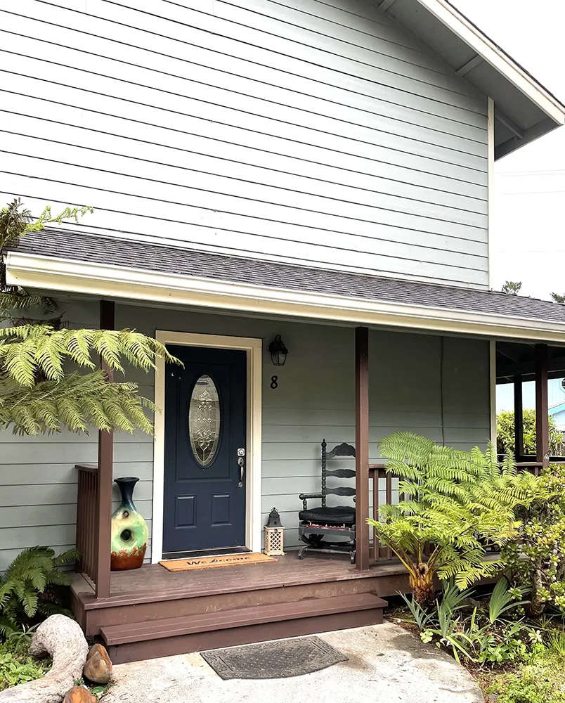 A cozy front porch of a light gray Humboldt house with a dark blue door, perfect for an Air BnB stay. The porch features wooden steps and posts, a rocking chair, and lush greenery, including ferns. An oval window in the door adds a charming touch to the inviting entrance.