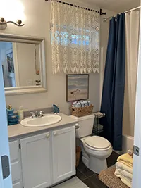 A small bathroom in this charming Humboldt Air BnB features a white vanity and sink, a toilet, and a bathtub with a dark shower curtain. An ornate lace curtain covers the window above the toilet, while a beach scene painting adorns the wall. Towels are neatly stacked on a bench.