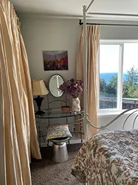 A cozy bedroom in this charming Air BnB boasts a large window offering an ocean view. The room features a white four-poster bed with a floral bedspread, a glass vanity table with a round mirror, and decorative items. Beige curtains frame the window, letting in natural light—perfect for your Humboldt getaway.