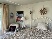 A cozy bedroom in this Humboldt Air BnB features a floral-patterned bed with a white metal frame and a large floral wreath on the wall above it. Beside the bed is a plush armchair with a dark cushion, complemented by a white lamp and nightstand. A painting and mirror decorate the wall, completing the charming atmosphere.