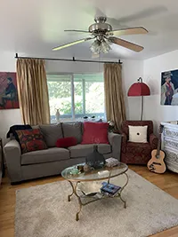 A cozy living room in a charming Air BnB features a gray sofa with colorful cushions, a red armchair, a glass coffee table, and a beige rug. A guitar sits beside the armchair. The walls showcase Humboldt-inspired artwork, and golden curtains on a large window let in natural light. A ceiling fan is above.