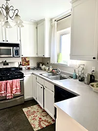 A bright Air BnB kitchen with white cabinets, countertops, and appliances. A window with white curtains is above the sink. A stove and microwave are on the left, with red and pink dish towels hanging from the oven handle. A colorful rug is placed on the floor, reflecting the charm of Humboldt.