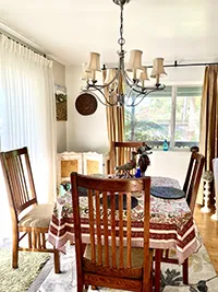 A cozy dining room with a wooden table and four chairs. The table is covered with a patterned tablecloth and set with blue placemats. A chandelier with candle-like bulbs hangs above. Sunlight streams in through large windows with sheer and golden curtains, offering a perfect Air BnB experience in Humboldt.