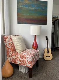 A cozy corner in this Humboldt Air BnB features a chair with orange floral upholstery, a white pillow, a colorful lamp with a white shade, and an acoustic guitar placed against the wall. Above the chair hangs an abstract painting with various shades of blue, green, and brown.