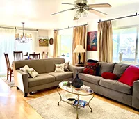 A bright living room in this charming AirBnB features two grey sofas adorned with colorful cushions, a glass coffee table with a decorative item, a rug, and several lamps. Large windows with beige curtains allow natural light to fill the room. A dining area with a chandelier is visible in the background.