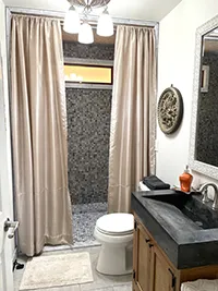 This modern Humboldt Air BnB bathroom features a shower area enclosed by beige curtains, a white toilet next to a wooden vanity with a dark countertop and sink, and a large mirror with a decorative frame above it. The lighting fixture includes three elegant lights.