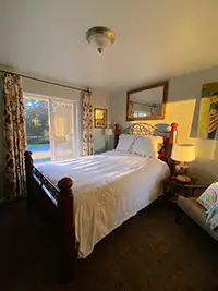 A cozy AirBnB bedroom in the heart of Humboldt, featuring a wooden bedframe and white bedding, illuminated by warm natural light from a glass sliding door on the left. The room boasts floral curtains, a bedside table with a lamp, a decorative mirror above the bed, and a cushioned chair with a colorful pillow.