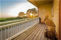 Wooden balcony with rustic chairs overlooking coastal grassland and trees at sunset.