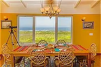 Dining table with wicker chairs and colorful placemats in front of large windows overlooking the Pacific Ocean.