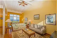 Bright living room with floral and gray sofas, wooden coffee table, vaulted ceiling, and ocean views in the background.