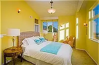 Bright yellow bedroom with a queen bed, wicker chair, and large windows offering coastal views.